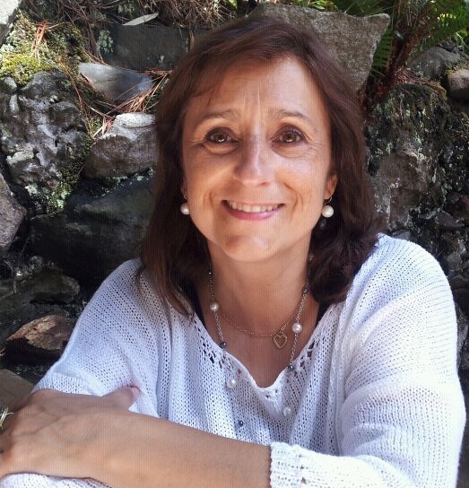 Mujer sonriente con cabello castaño, collar y suéter blanco, posando frente a rocas y vegetación.